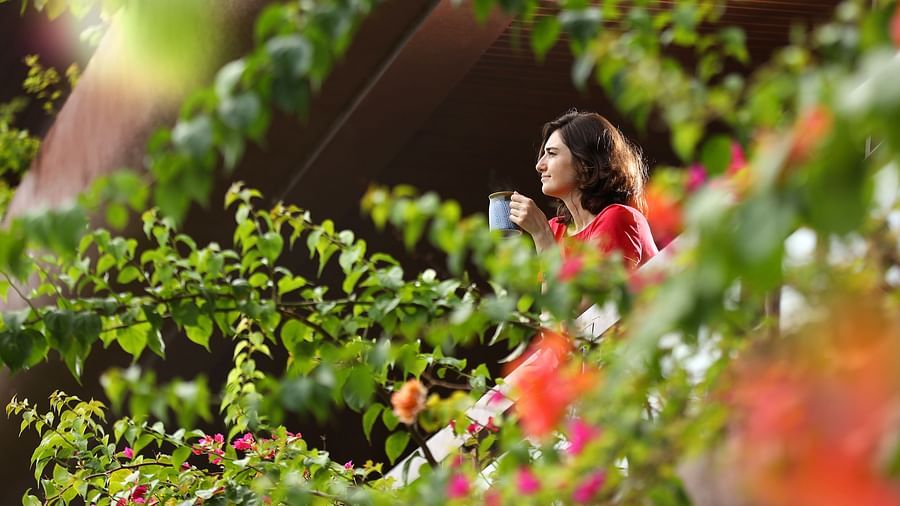 A woman seen amidst the green surroundings standing in the balcony with a mug of hot beverage and enjoying the nature at Uday Backwater Resort