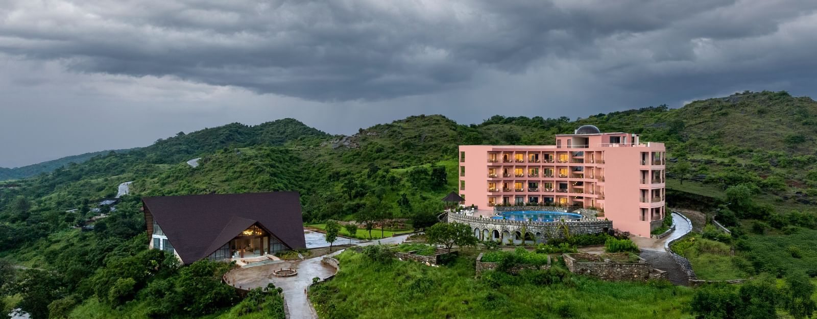 A view of the Osian Valley Resort & Spa showing the a pink multi-storey building, a separate dark-roofed building, and the surrounding green, hilly landscape under a cloudy sky.
