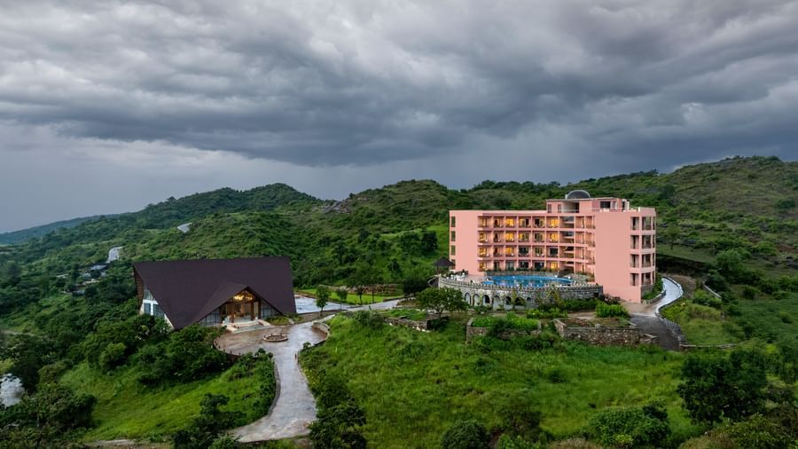 A view of the Osian Valley Resort & Spa showing the a pink multi-storey building, a separate dark-roofed building, and the surrounding green, hilly landscape under a cloudy sky.