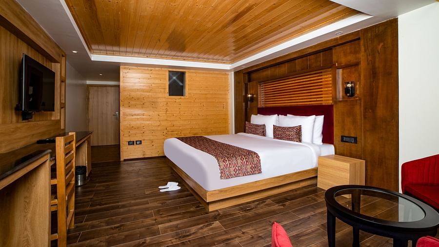 A hotel room with a large bed featuring white linens and red accents, wooden panelling on the walls and ceiling, dark wood flooring, a flat-screen TV mounted on the wall, a work desk with chair, and a red armchair with a round glass-top table - Yashshree L