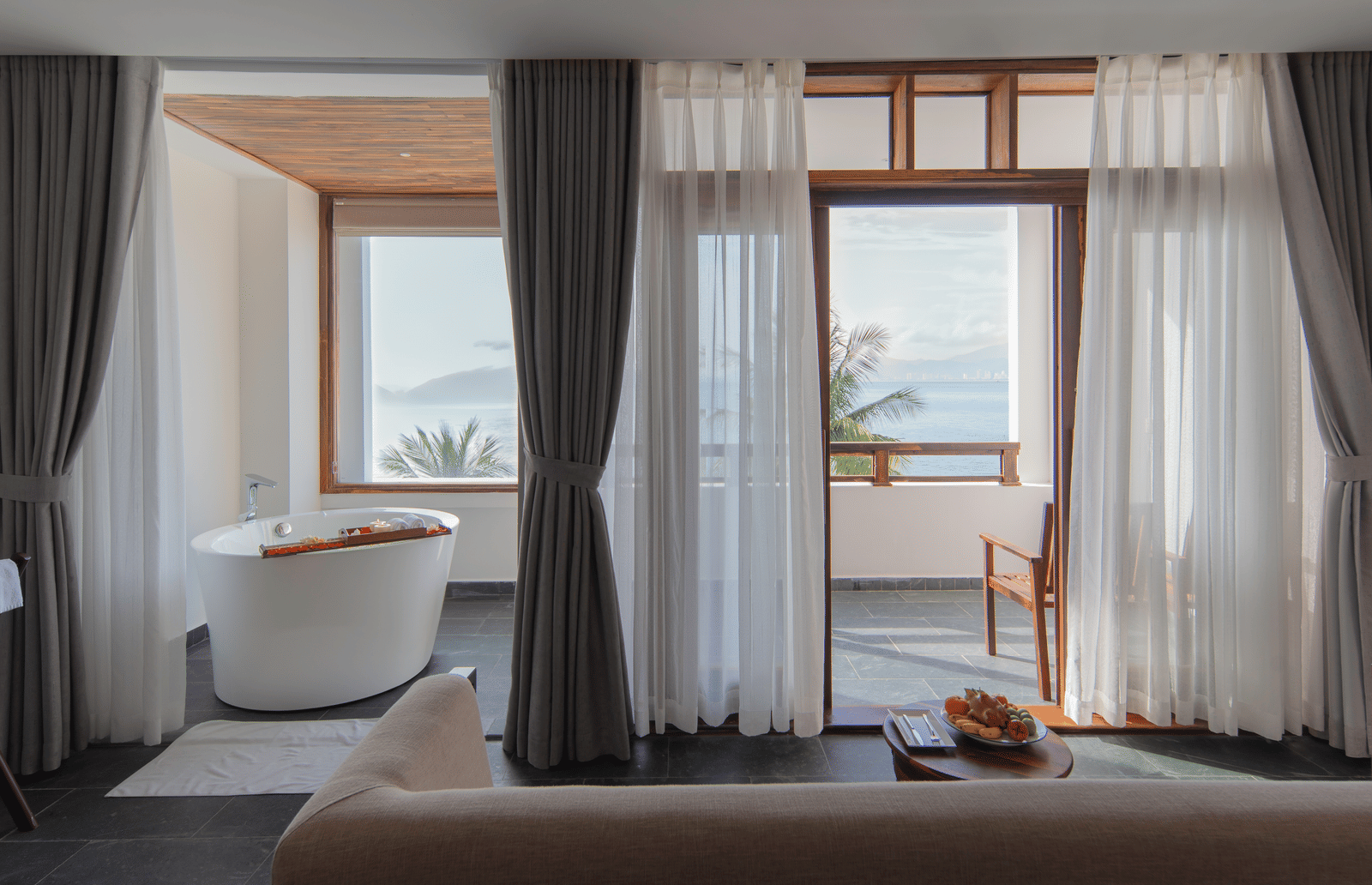 Executive Ocean Suite at Alibu Resort Nha Trang, Vietnam with bathtub, glass windows, seating area, open curtains and garden view bringing outside light.