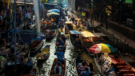 an overview of a floating market with different boats selling from fruits to hand bags and shops lined up on the buildings on either side of the waterbody. These floating markets are a must-visit during while on excursions from Bangkok.