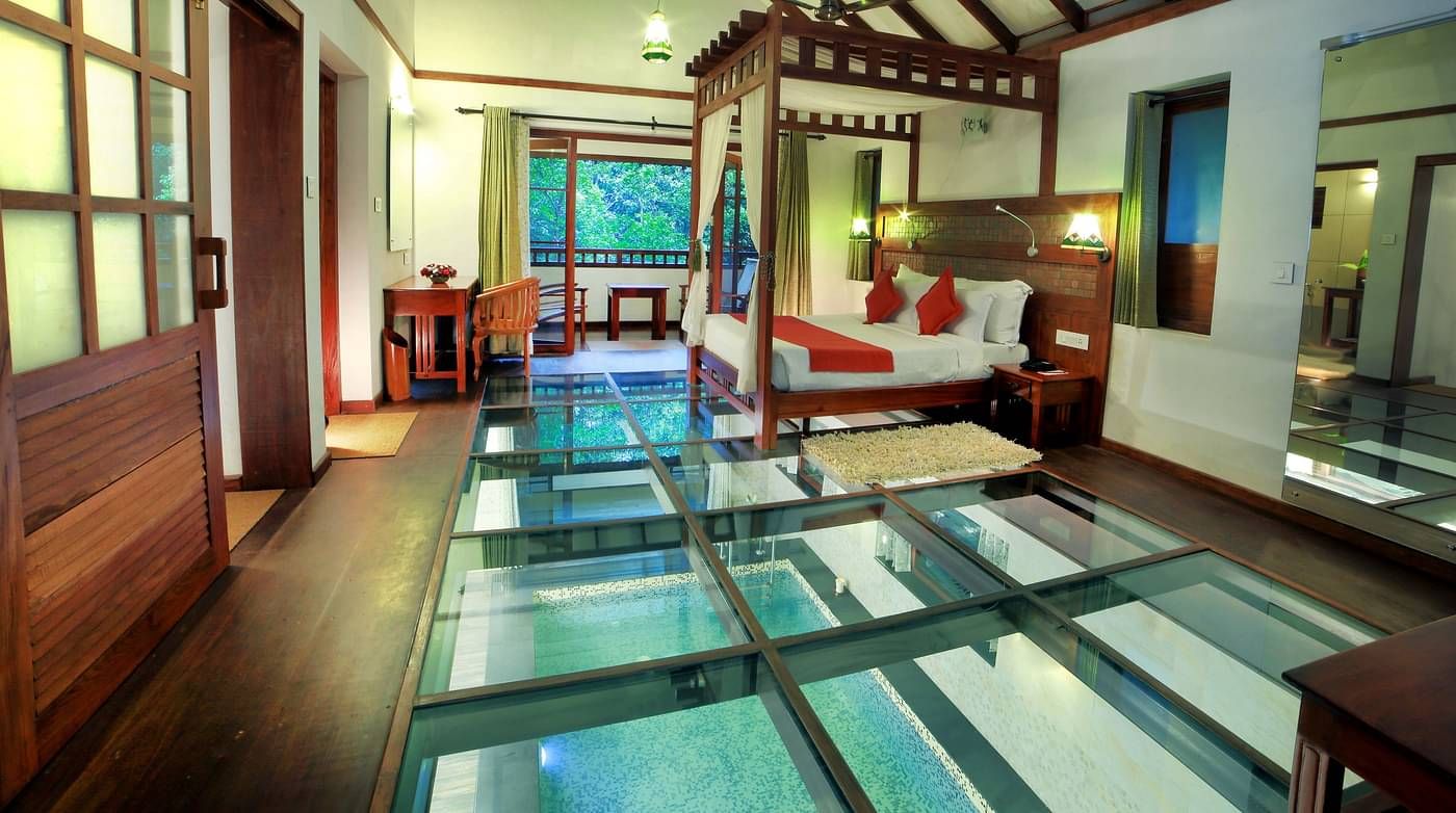 A side view of a glass-floored room with a bed and other wooden furnishings | Vythiri Resort. Wayanad