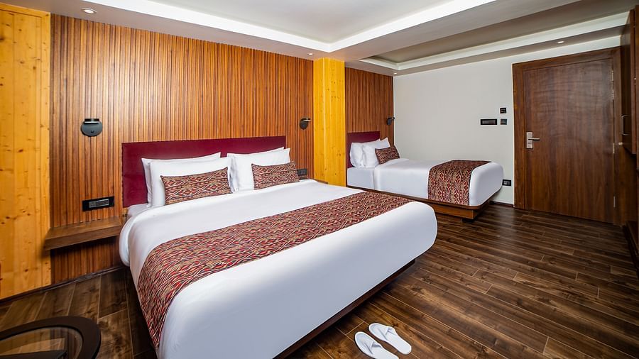Spacious hotel room with two beds—one queen and one single—featuring red headboards and patterned bed runners, wooden flooring, warm wood-panelled walls, and a closed wooden door in the background - Yashshree Lachen.