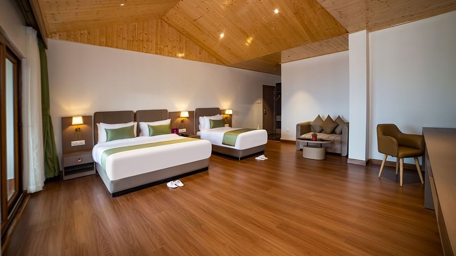 A hotel room with two neatly made beds featuring green accents, a cosy sitting area with armchairs, a wooden floor, and a large glass door opening to a balcony with scenic views - Yashshree Pelling Chekhim Retreat.