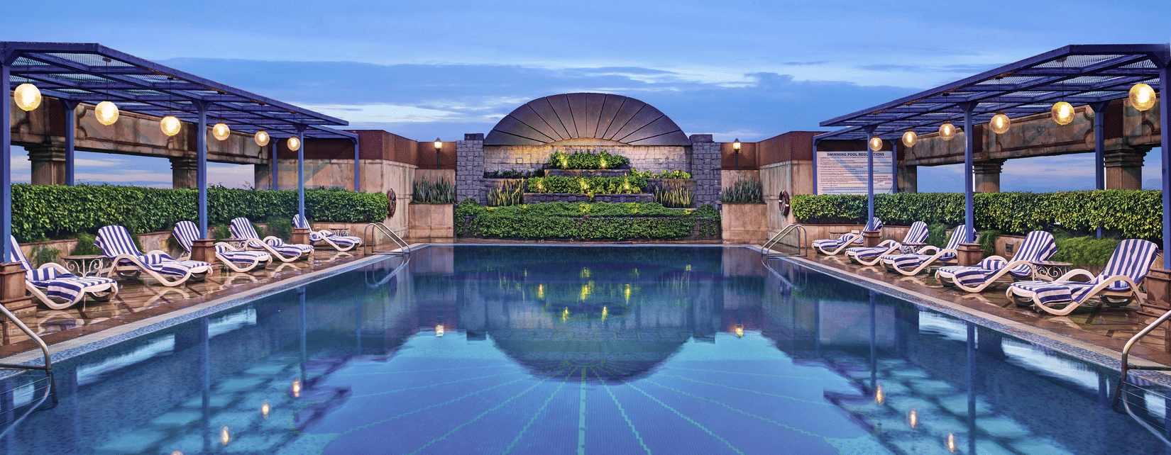 Extended view of the rooftop swimming pool at the best hotel for corporate outing in Gurgaon featuring lounge chairs with shades and ample lighting, and a blue sky -The Bristol Hotel, Gurgaon