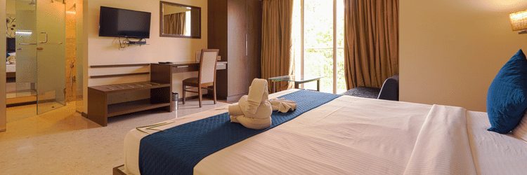 A room at Magnus Star Residency, Koregaon Park Annexe, featuring a neatly made bed with decorative folded towels, a desk with a chair, a mounted television, a wardrobe, a ceiling fan, a glass-walled bathroom entrance, and windows with curtains.