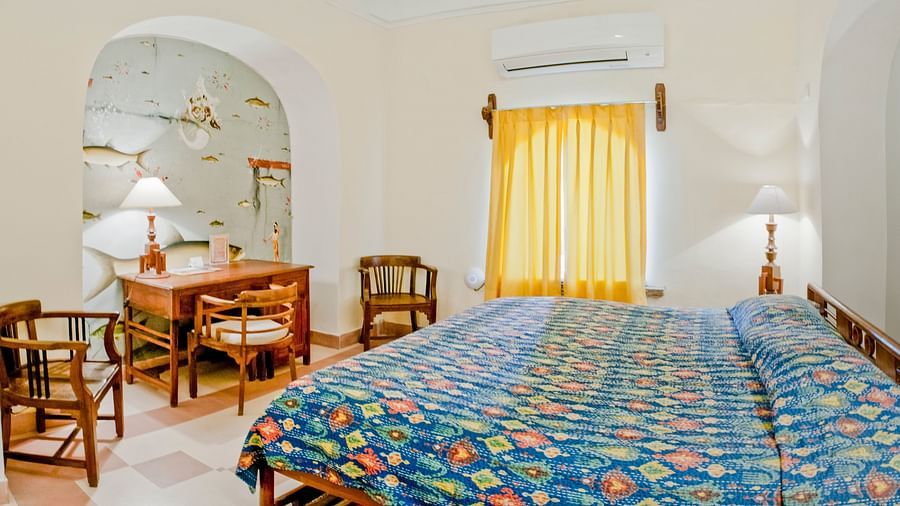 Bedroom with a bed featuring a blue patterned cover, bedside lamps, a desk and chairs in an alcove, yellow curtains, and AC at Tijara Fort-Palace, Alwar.