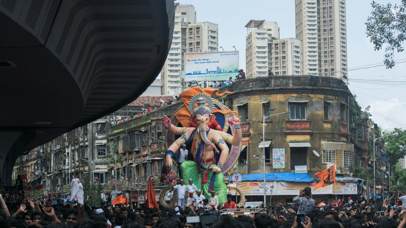 A vibrant Ganesh Chaturthi procession passes through a Mumbai street beneath cloudy monsoon skies.