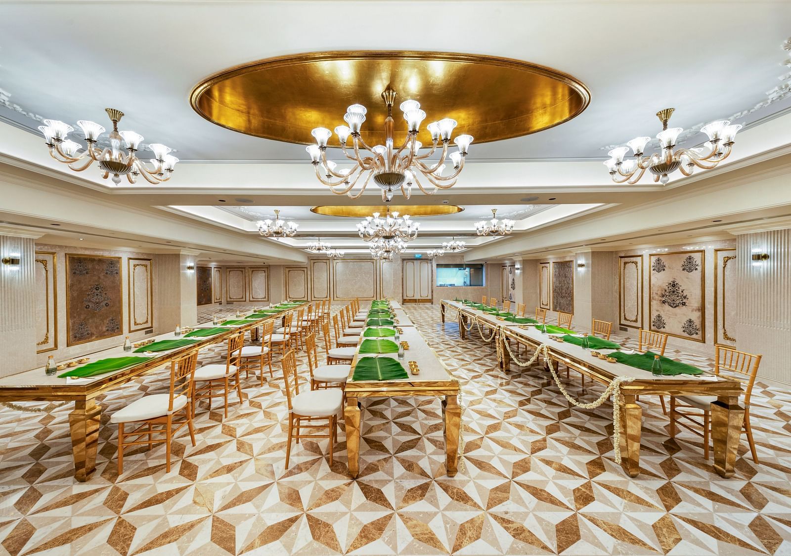 A luxurious dining hall at Grand Chennai by GRT Hotels  featuring multiple long tables with golden chairs, illuminated by ornate chandeliers.