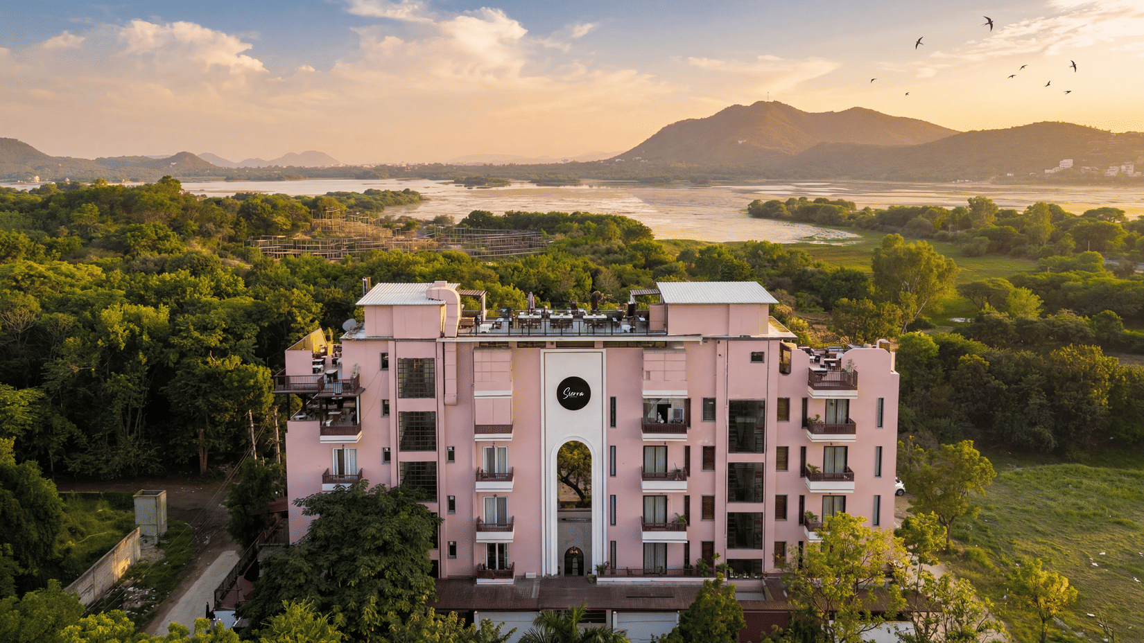An aerial view of a pink building surrounded by dense green trees, hills, a lake at The Sierra - By The Lake.