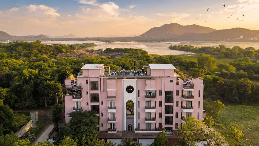 An aerial view of a pink building surrounded by dense green trees, hills, a lake at The Sierra - By The Lake.