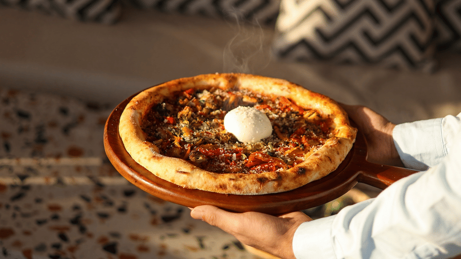 A person holds a wooden platter serving a freshly baked whole pizza topped with a ball of cheese at a dining table at The Sierra - By The Lake.