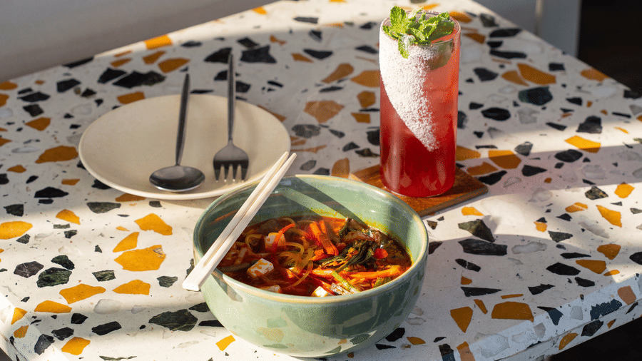 A bowl of noodles served with chopsticks sits next to a tall glass filled with a layered drink on a patterned tabletop at The Sierra - By The Lake.