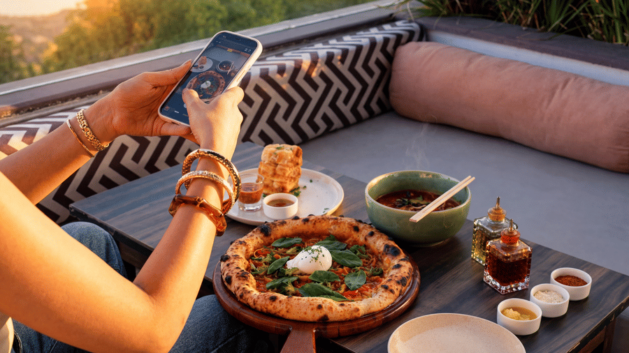 A person uses a smartphone to take a photo of a dining spread, including pizza, bowls, and condiments, on an outdoor sofa at The Sierra - By The Lake.