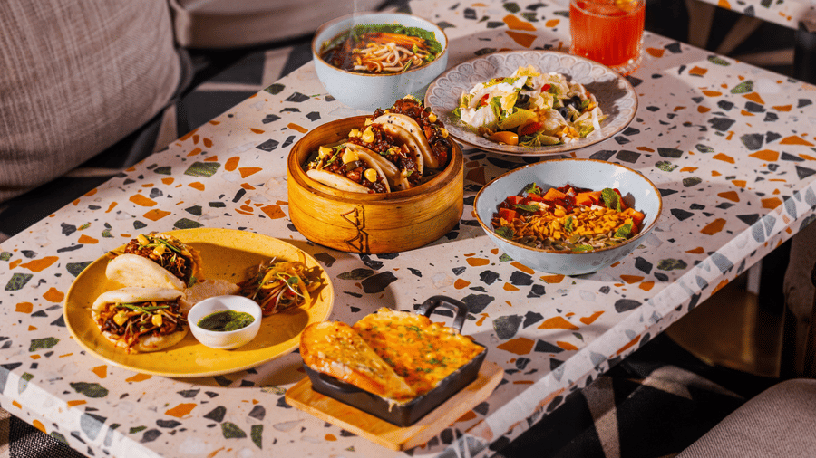 A diverse spread of Asian food dishes, including a dim sum basket, buns, and side bowls, is arranged on a patterned table at The Sierra - By The Lake.