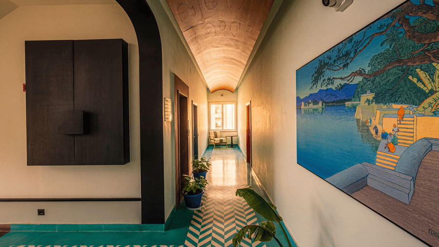 A well-lit indoor hallway displaying a large painting on the wall, doorways, plants at The Sierra - By The Lake.