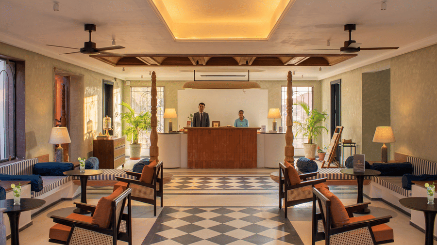 Two receptionists stand behind a wooden desk in the symmetrical lobby area of The Sierra - By The Lake, which features armchairs, ceiling fans, and tiled floors.