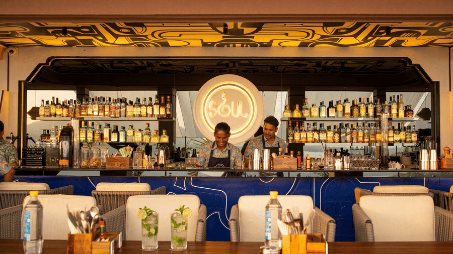 Two bartenders stand behind a fully stocked indoor bar counter at The Sierra - By The Lake, with shelves of bottles, glasses, and seating arranged in front.