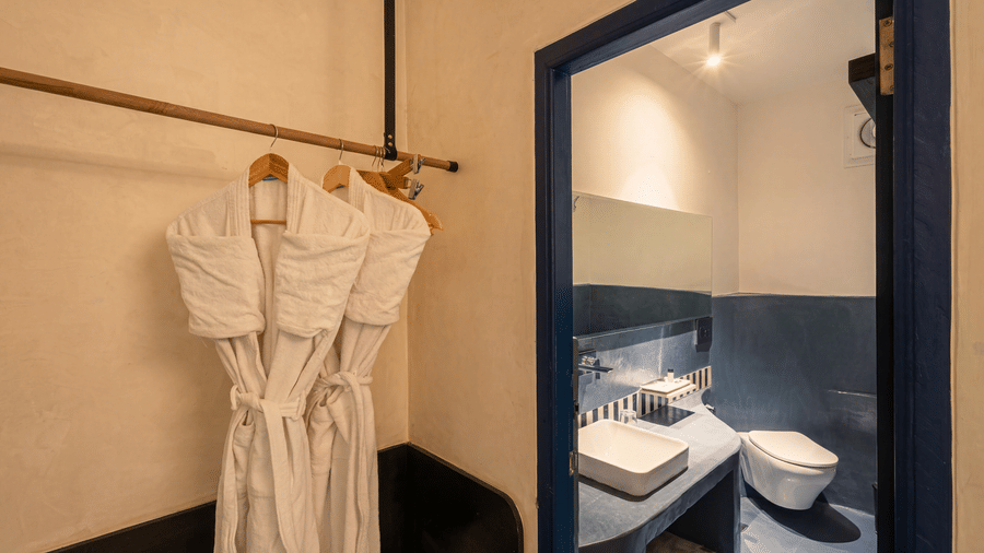 A dressing area with white bathrobes on hangers alongside a modern washbasin, mirror, and toilet at The Sierra - By The Lake.