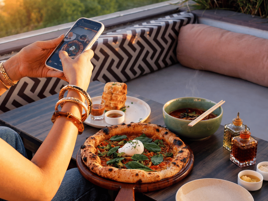 A person uses a smartphone to take a photo of a dining spread, including pizza, bowls, and condiments, on an outdoor sofa at The Sierra - By The Lake.