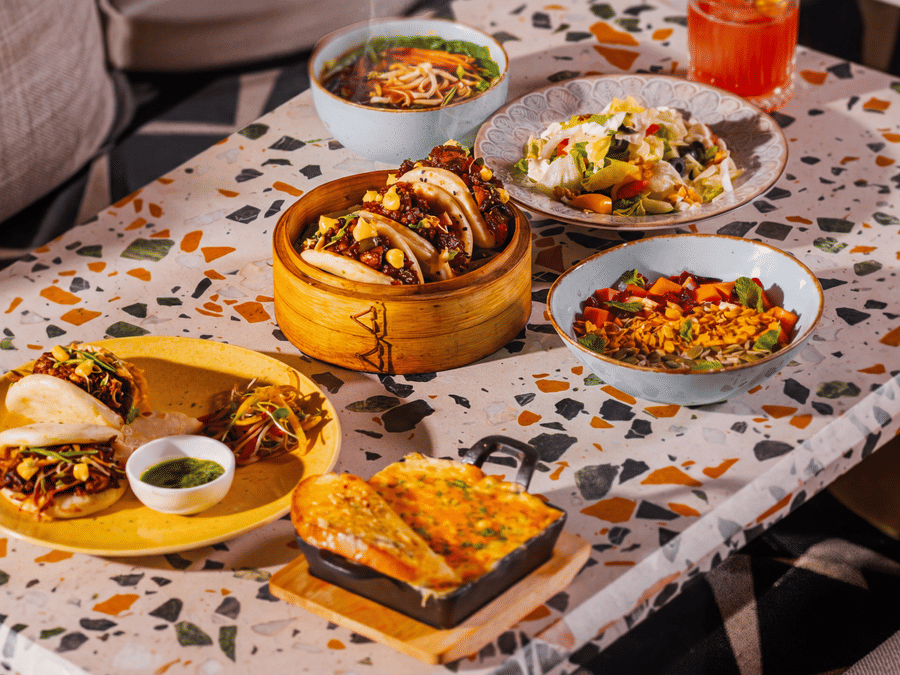 A diverse spread of Asian food dishes, including a dim sum basket, buns, and side bowls, is arranged on a patterned table at The Sierra - By The Lake.