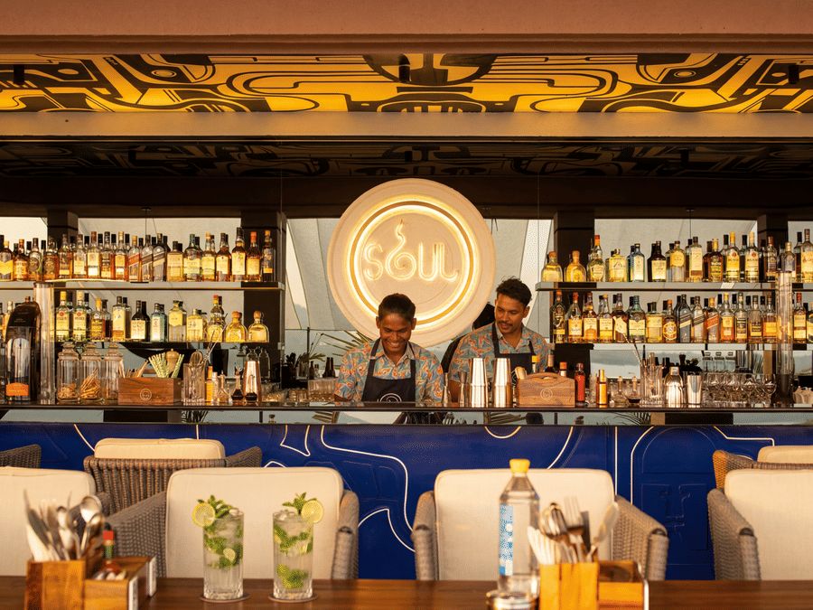 Two bartenders stand behind a fully stocked indoor bar counter at The Sierra - By The Lake, with shelves of bottles, glasses, and seating arranged in front.