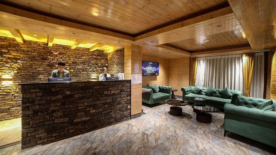 Brightly lit hotel lobby with a stone reception counter, light wooden ceiling, green cushioned seating in a cosy lounge area, and a wall-mounted TV adding a modern touch - Yashshree Lachen.
