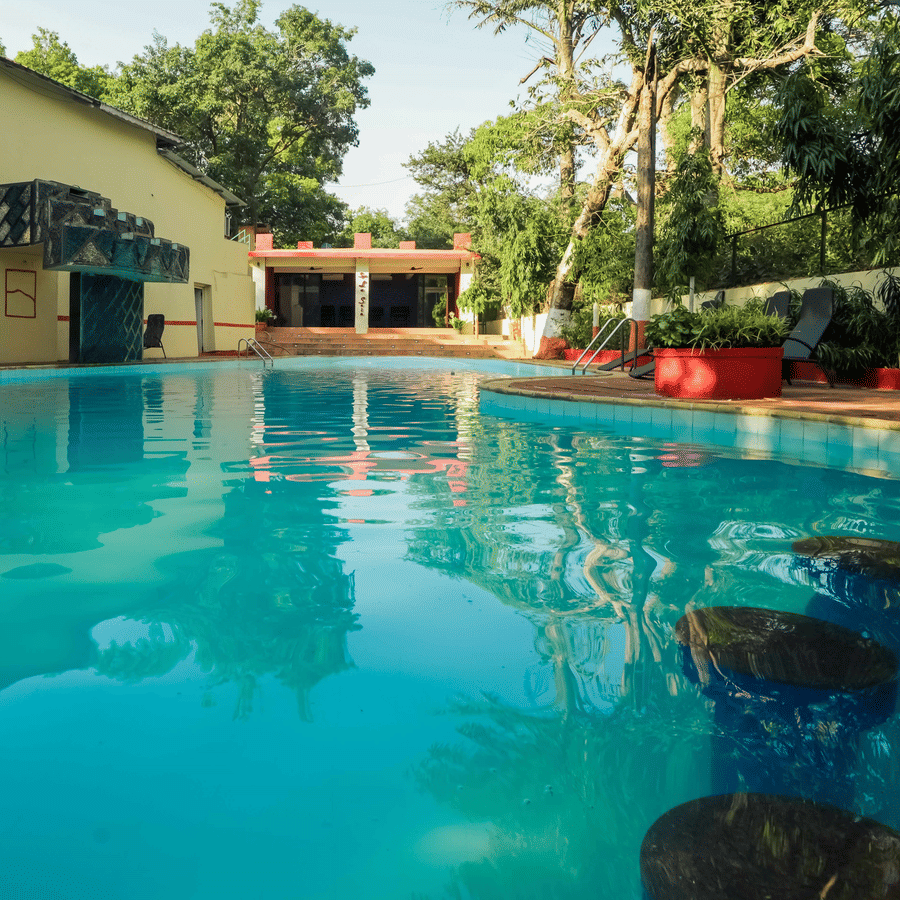 A serene outdoor view at Adamo The Village featuring a swimming pool, a cottage, and greenery in the background.
