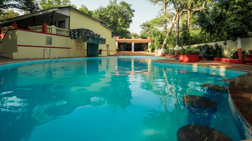 A serene outdoor view at Adamo The Village featuring a swimming pool, a cottage, and greenery in the background.