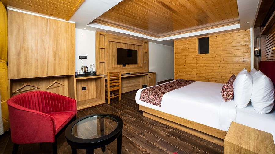 Stylish hotel room featuring a large bed with white linens and red accents, wooden walls and ceiling, dark wooden flooring, a red upholstered chair with a glass-top coffee table, and a wall-mounted TV with a built-in wooden desk and storage unit - Yashshre