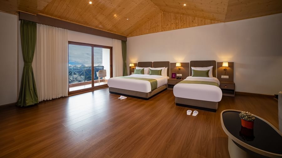 Spacious hotel room with two beds, wooden flooring, a large glass door leading to a balcony with mountain views, and cosy modern furnishings under a wooden ceiling - Yashshree Pelling Chekhim Retreat.