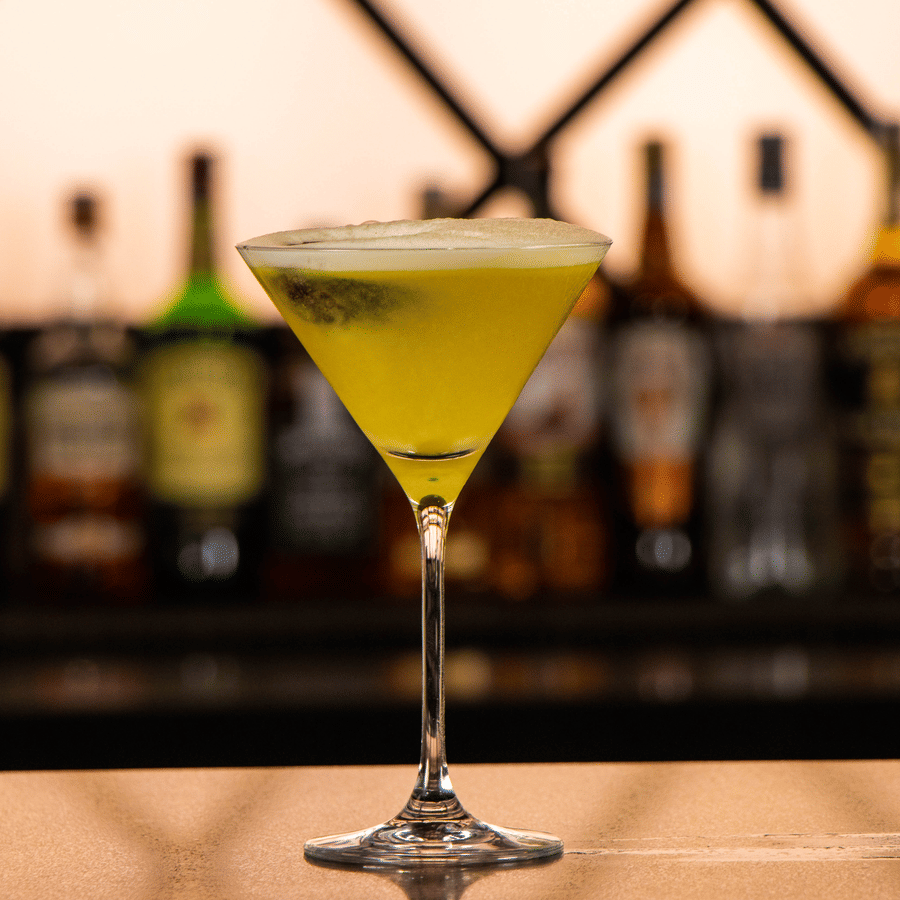 Cocktail glass with a green drink placed on a bar counter, bottles lined up in the background at Asiatic Rooftop Bar and Restaurant.