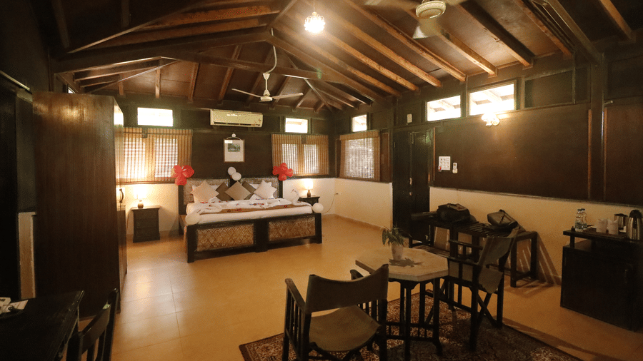 A bedroom with a bed, 2 armchairs, a center table, and a wooden ceiling with exposed beams at Infinity Resorts Bandhavgarh.