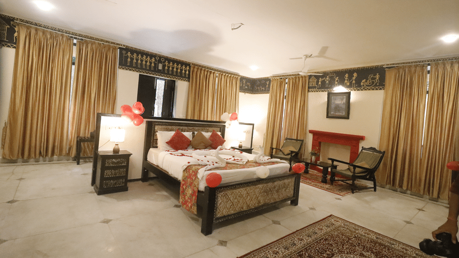 A bedroom with a large bed, sofa seating, side tables, lamps, and wide curtained windows at Infinity Resorts Bandhavgarh.