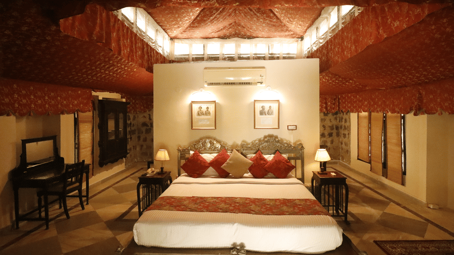 The bedroom with a wooden ceiling, double bed, side seating chairs, and symmetrical layout with lamps at Infinity Resorts Bandhavgarh.