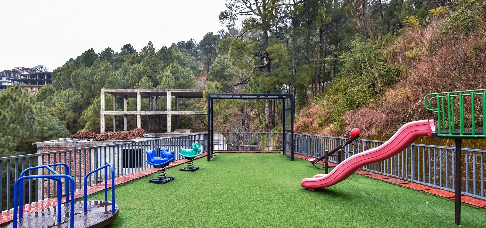 A children's play area with colourful slides and climbing structures, set on a well-maintained grassy surface. The space is surrounded by lush greenery and provides a safe, fun environment for kids.