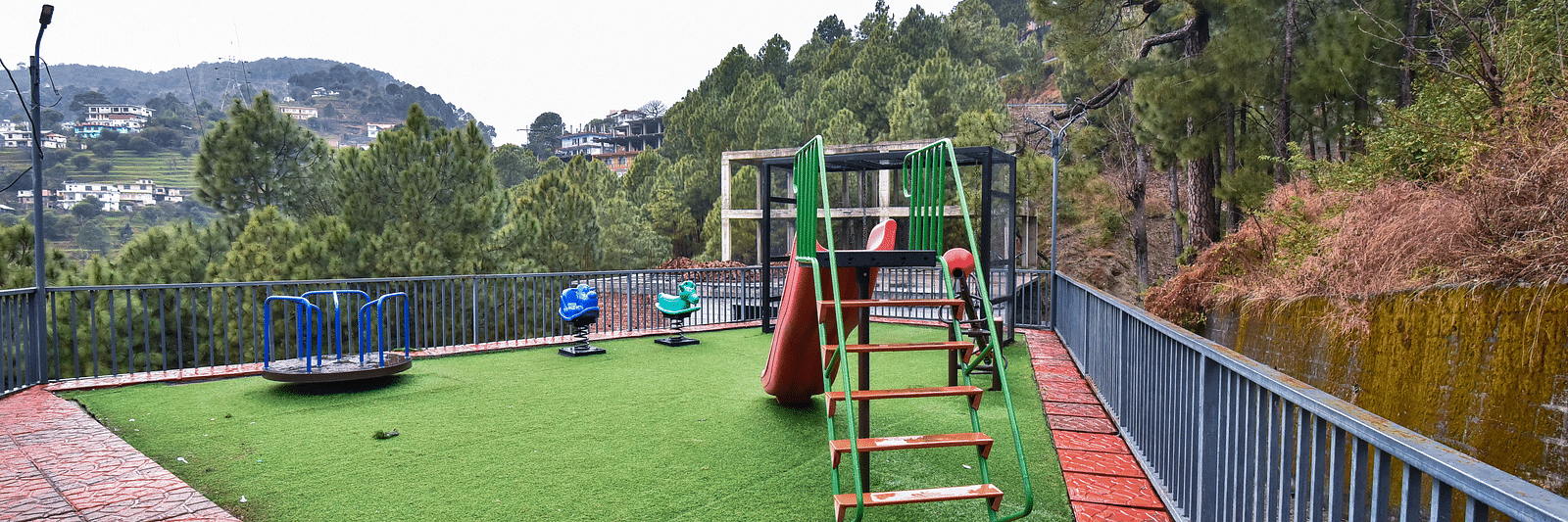A children's play area with a slide and colourful play equipment on a well-maintained grassy surface, surrounded by a fence and beautiful views.