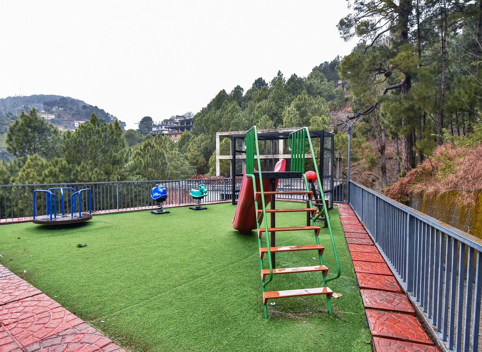 A children's play area with a slide and colourful play equipment on a well-maintained grassy surface, surrounded by a fence and beautiful views.
