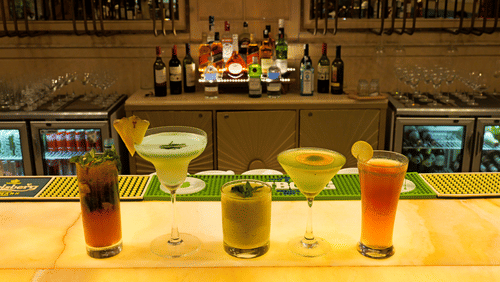 A close-up view of a bar counter with several colorful cocktails in glasses. Bottles are arranged on a shelf behind the bar, and a refrigerated display case is also visible - The Gaurs Sarovar Premiere