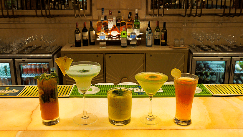 A close-up view of a bar counter with several colorful cocktails in glasses. Bottles are arranged on a shelf behind the bar, and a refrigerated display case is also visible - The Gaurs Sarovar Premiere