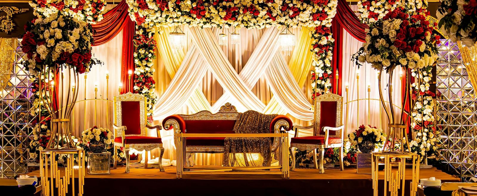 A beautifully decorated stage with flowers and drapes featuring a grand couch in the centre stage with a brightly lit background.