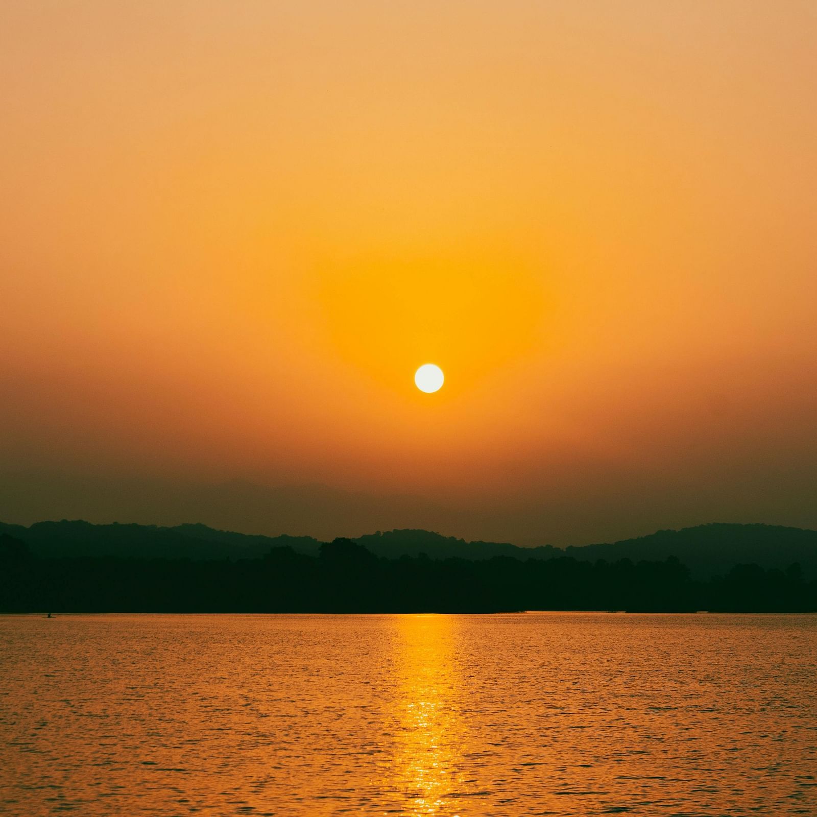 A sunrise over Sukhna Lake, with the sun appearing as a bright orb reflecting on the water against a backdrop of dark hills and a gradient sky. 