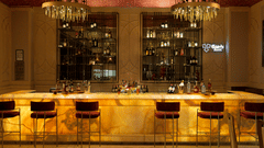 A wider view of a bar with a glowing countertop and red bar stools. Illuminated shelves displaying bottles line the back wall. Decorative chandeliers hang above the bar - The Gaurs Sarovar Premiere