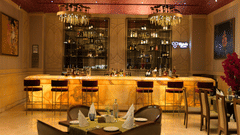A dimly lit bar area with a glowing countertop and several bar stools. Shelves stocked with bottles are visible in the background, illuminated from within. A few tables with chairs are situated near the bar - The Gaurs Sarovar Premiere