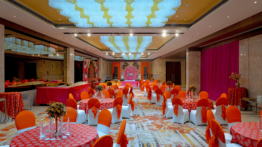 A large indoor venue at The Gaurs Sarovar Premiere, featuring round tables and chairs arranged for a banquet or reception, and ornate lighting.