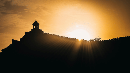 A dramatic silhouette of a fortress wall and a watchtower on a hilltop, with bright sunbeams bursting from behind the structure.