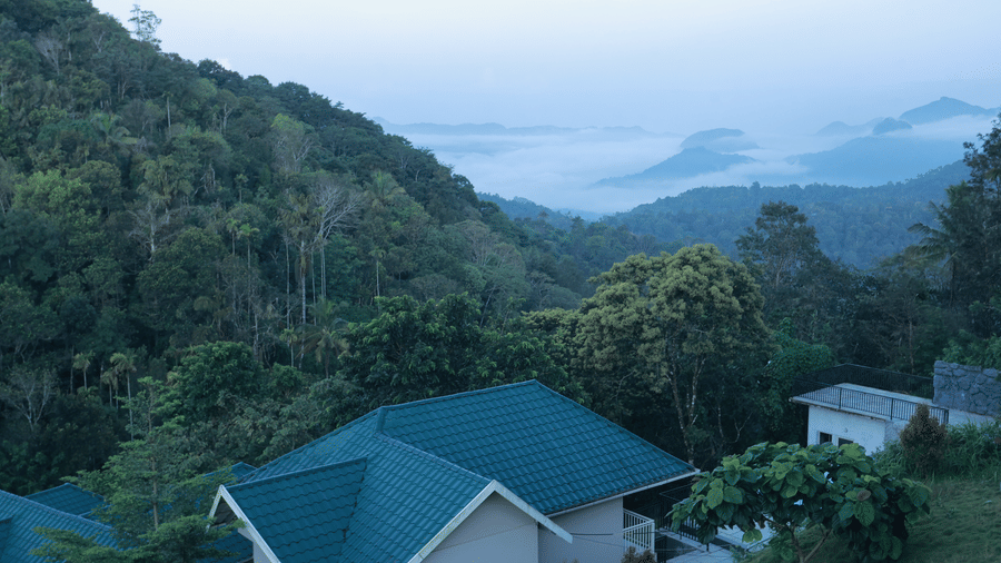 A slope of a hill covered with a thick green cover overlooking the villas at Aveda Mountains and Mist Resort and Spa, Munnar, alongside the mountain valleys covered with a thick cloud cover.