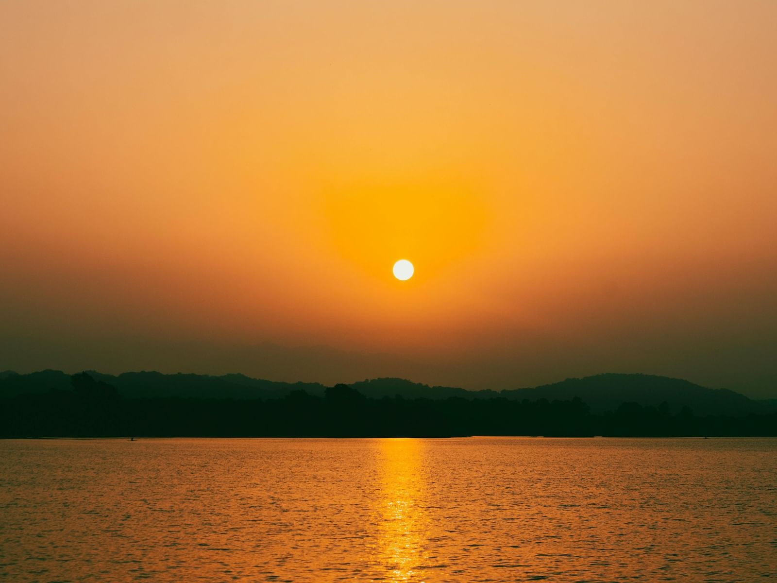 A sunrise over Sukhna Lake, with the sun appearing as a bright orb reflecting on the water against a backdrop of dark hills and a gradient sky. 