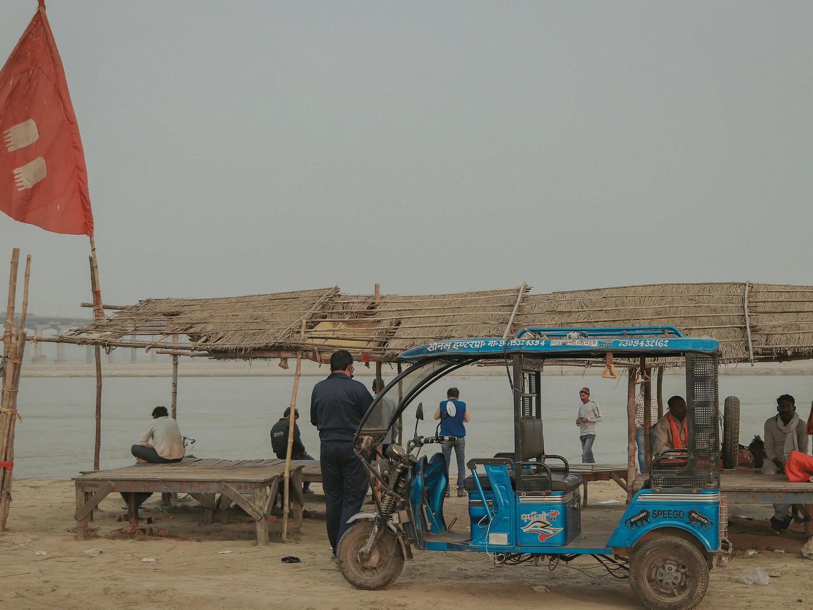 A rickshaw parked near a riverside settlement with bamboo huts and red flags in the background.